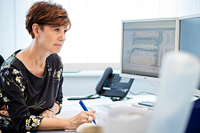 Eine Mitarbeiterin mit kurzem, dunklem Haar und dunkel-gemustertem Oberteil arbeitet an ihrem Schreibtisch und schaut dabei auf Ihre Computer-Monitore, Bild: ASV