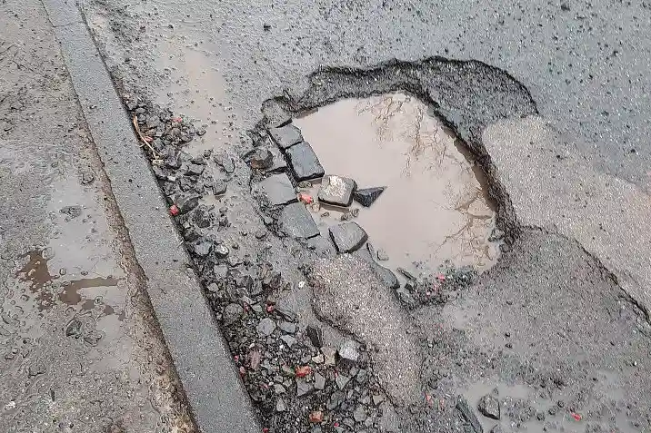 Ausschnitt einer feuchten Straße mit großem, Regenwasser gefülltem Schlagloch. Links vom Schlagloch sind unterschiedlich große und kleine Steine sichtbar. Bild: ASV