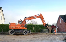 Eine Straßenbaustelle mit Bagger und Bauarbeiter, im Hintergrund zwei Häuser