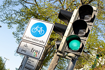 Nahaufnahme von einer grünen Ampel und einem Straßenschild Fahrradstraße. Im Hintergrund ein grüner Baum vor blauem Himmel, Bild: ASV