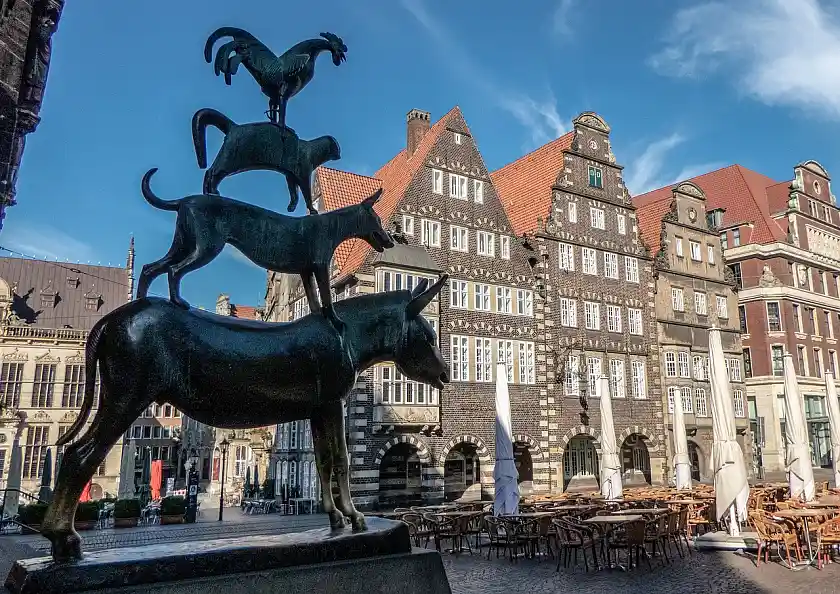 Links Statue der Bremer Stadtmusikanten vor blauem Himmel. Im Hintergrund historische Gebäude und Teile des Marktplatzes. Rechts auf dem Bild Stühle, Tische und Sonnenschirme