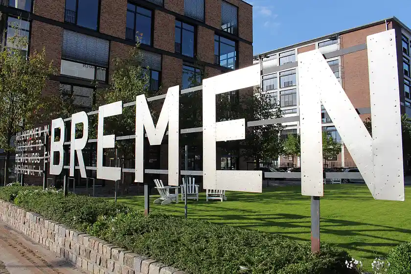 Schriftzug Bremen in großen Metall Lettern über einer grünen Wiese. Im Hintergrund Gebäude mit rötlichen Ziegeln