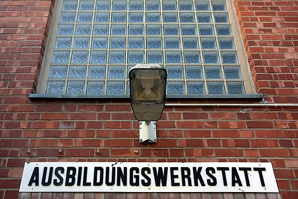 Blick auf einen Ausschnitt einer roten Ziegelwand. Oben sind viele Glasbausteine als Fenster, darunter eine einzelne Lampe. Unter der Lampe ein Schild mit der Aufschrift Ausbildungswerkstatt