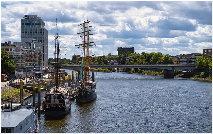 Blick auf die Weser, die von einer Brücke überspannt wird. Linkerhand befinden sich Segelschiffe und Fußgänger auf der Promenade. Es sind Gebäude und grüne Bäume sichtbar