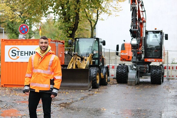 Ein lächelnder junger Mann mit Bart in orangefarbener Sicherheitsjacke vor einem Container und Baumaschinen