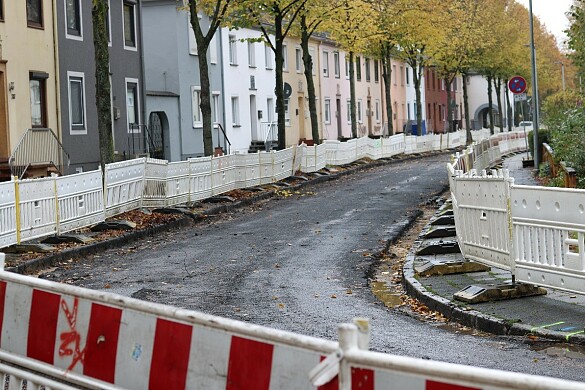 Ein eingezäunter Straßenabschnitt, die Deckschicht ist bereits abgefräst, links sind mehrere grüne Bäume und Reihenhäuser sichtaber