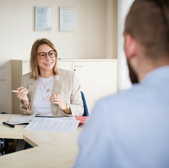 Eine lächelnde, blonde Frau mit Brille  an einem Tisch mit einer zweiten, männlichen Person,  im Gespräch.