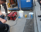 Vor Ort wird der Werkstoff aus Gummigranulat, Wasser und Harz angemischt in einem Eimer von einem dunkelhaarigen Mann. Im Hintergrund ein halboffener blauer Container, diverse Eimer, ein kleines, rotes Elektrogerät