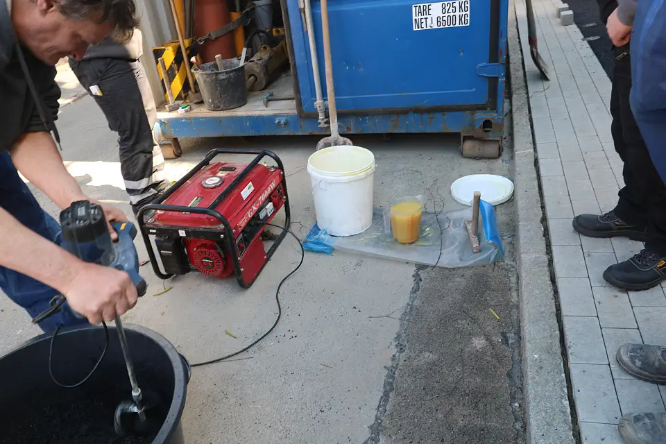 Vor Ort wird der Werkstoff aus Gummigranulat, Wasser und Harz angemischt in einem Eimer von einem dunkelhaarigen Mann. Im Hintergrund ein halboffener blauer Container, diverse Eimer, ein kleines, rotes Elektroger&auml;t