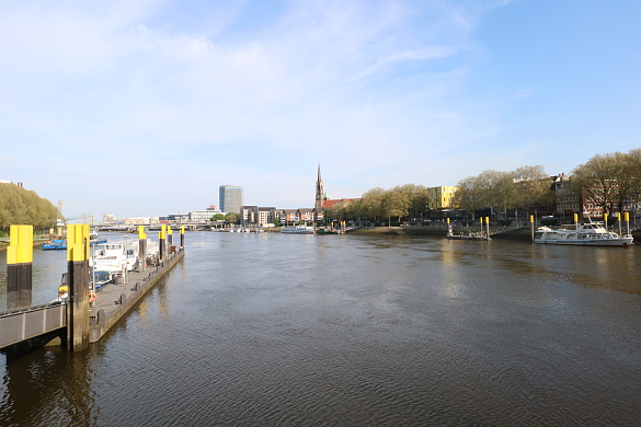 Blick auf die Weser aus dem Büro im Brückenkopf. Links  befindet sich ein Bootsanleger, rechts Schiffe. Beidseitig am Ufer grüne Bäume vor blauem Himmel, rechts sind Gebäude und ein Kirchturm