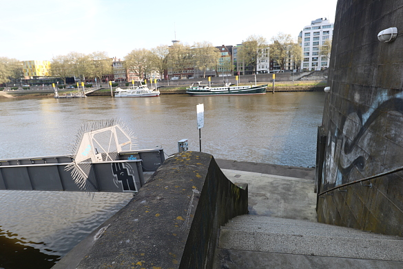 Treppenaufgang zum Brückenkopf und Blick auf die Weser und angelegte Schiffe am gegenüberliegendem Ufer. Dahinter die Weserpromenade mit grünen Bäumen, dahinter mehrere Gebäude 