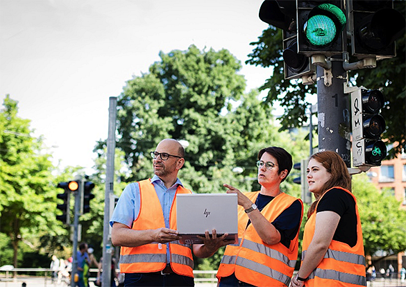 Drei Personen mit Warnweste vor einer Ampel. Planen an einem Labtop