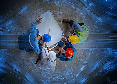 Blick von oben auf vier Bauingenieur*innen am Tisch, die auf ein großes Blatt schauen. Alle tragen Sicherheitshelme in unterschiedlichen Farben