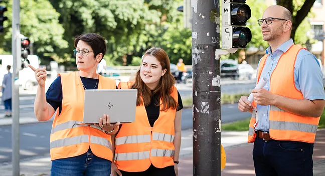 3 Mitarbeiter*innen mit orangen Westen vor einer Amppel